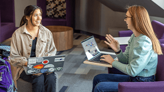 two girls sit on purple chairs in building lobby smiling and talking to each other while working on laptops